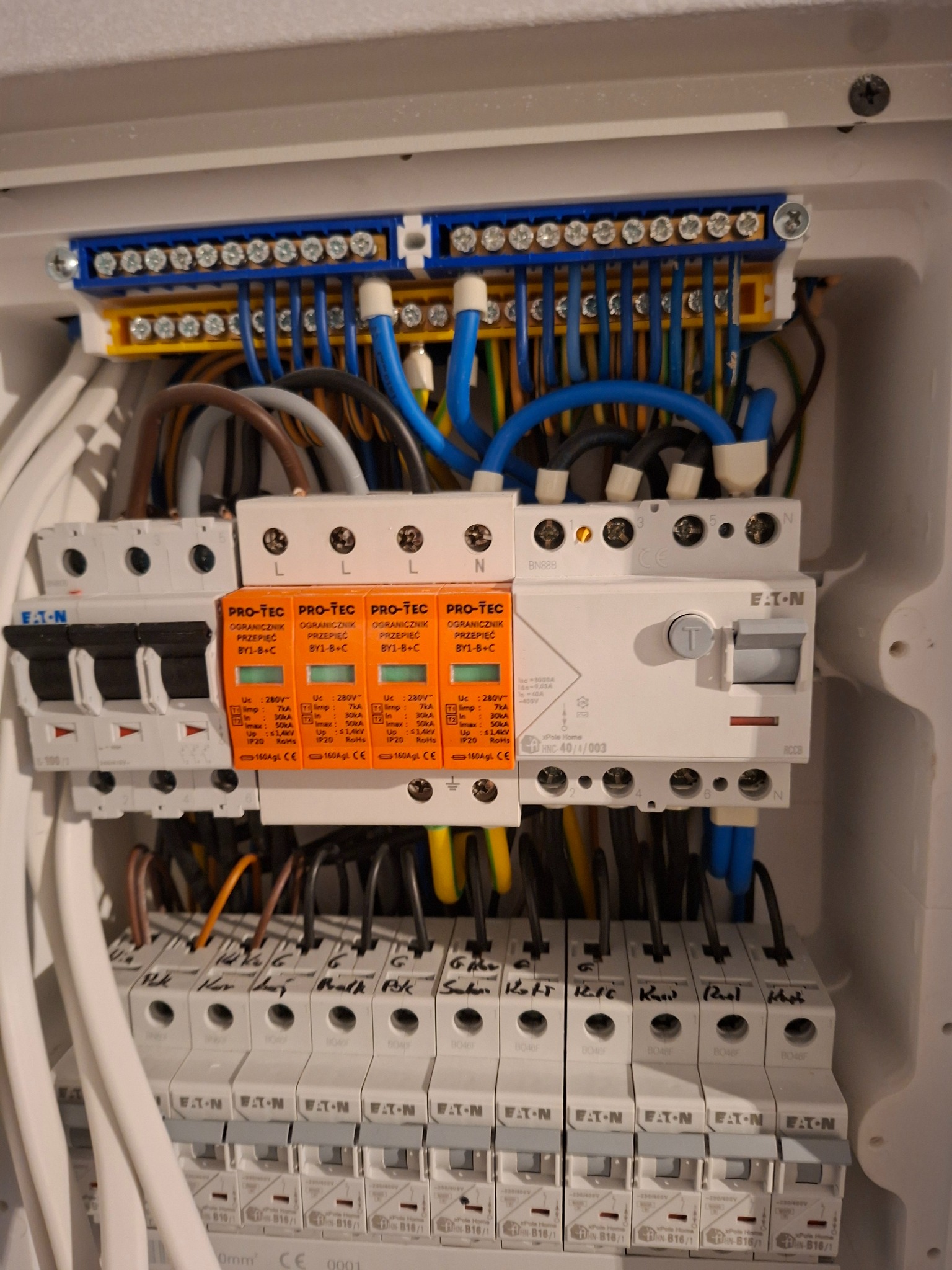 Rozdzielnica elektryczna z zabezpieczeniami przepięciowymi Pro-Tec i wyłącznikami Eaton, widoczne przewody w kolorach niebieskim, żółtym i brązowym.