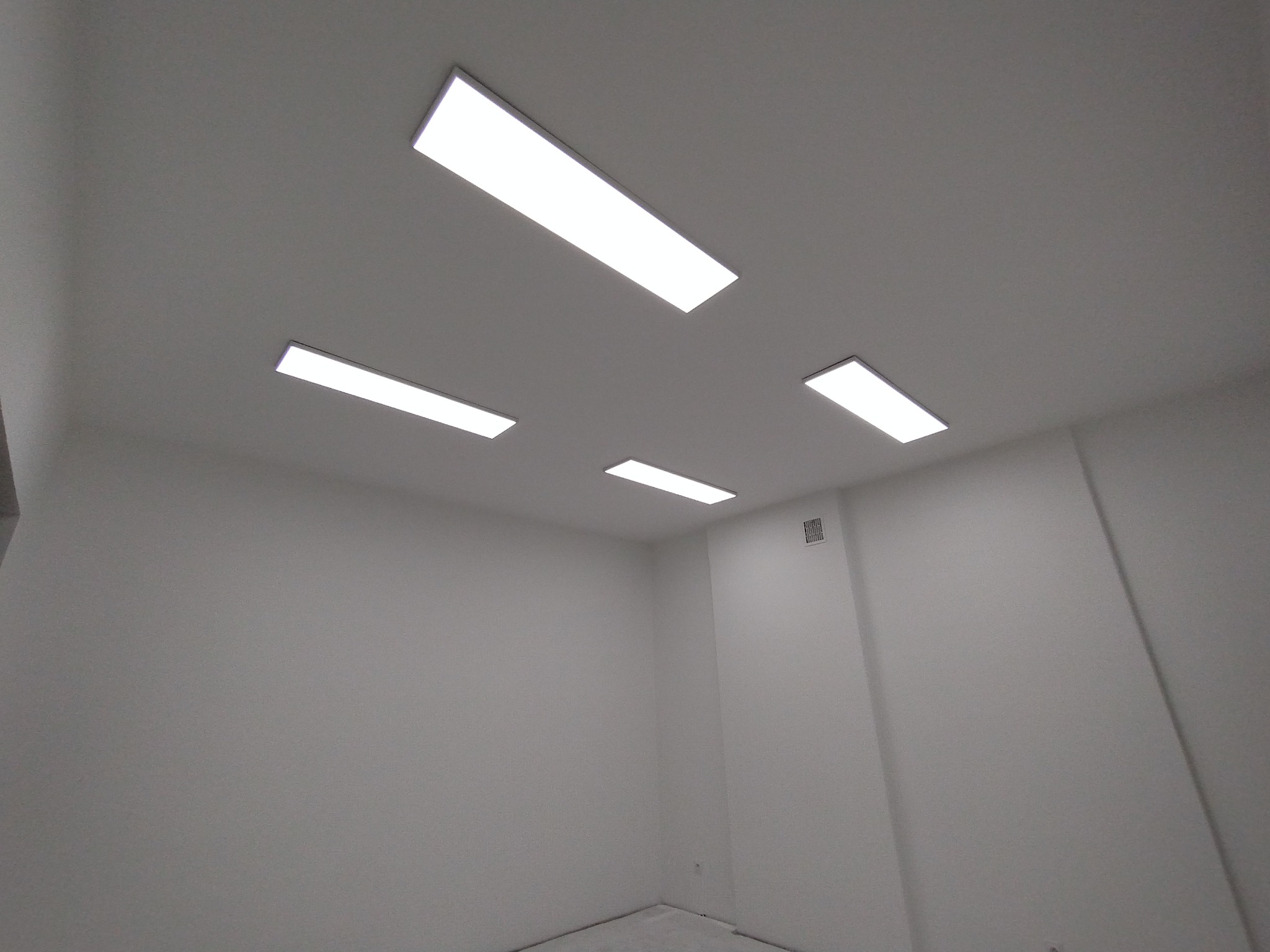 Białe, prostokątne panele LED w suficie, oświetlające puste, białe pomieszczenie. Widok pod kątem, ukazujący rozmieszczenie lamp i minimalistyczny design wnętrza.