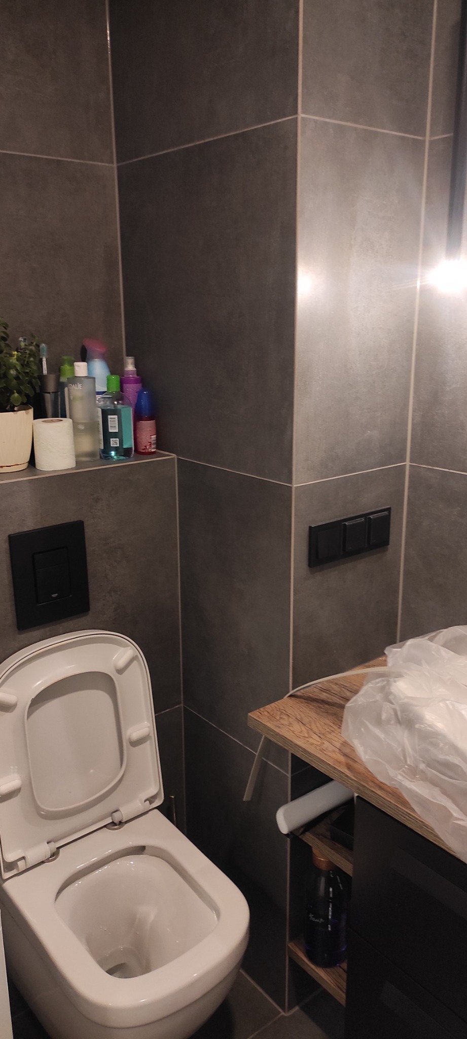 Nowoczesna toaleta wisząca w łazience z szarymi płytkami, widoczny fragment blatu z umywalką i kosmetyki na półce, czarne przyciski spłuczki i włączniki światła.