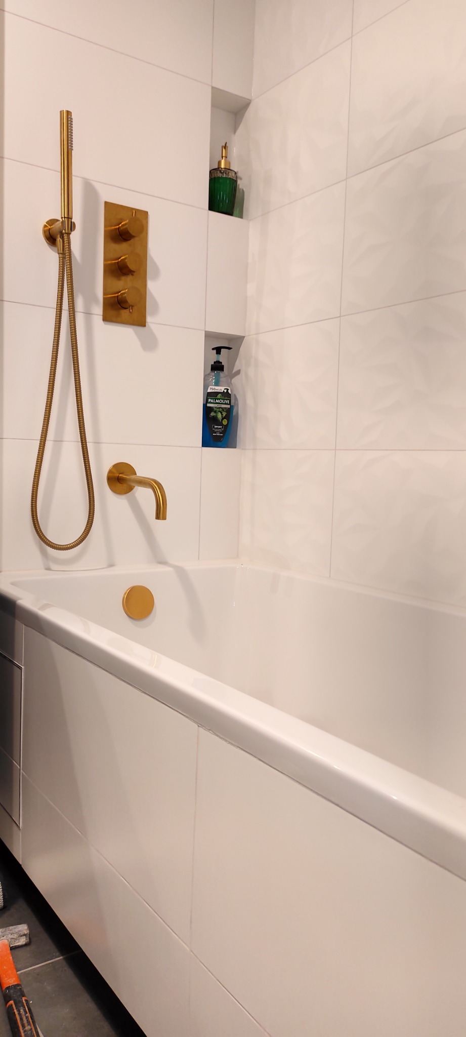 Biała wanna z baterią i prysznicem w odcieniu szczotkowanego złota na tle białych, teksturowanych płytek. Wnęka w ścianie z zielonym flakonem perfum i butelką mydła w płynie Palmolive.