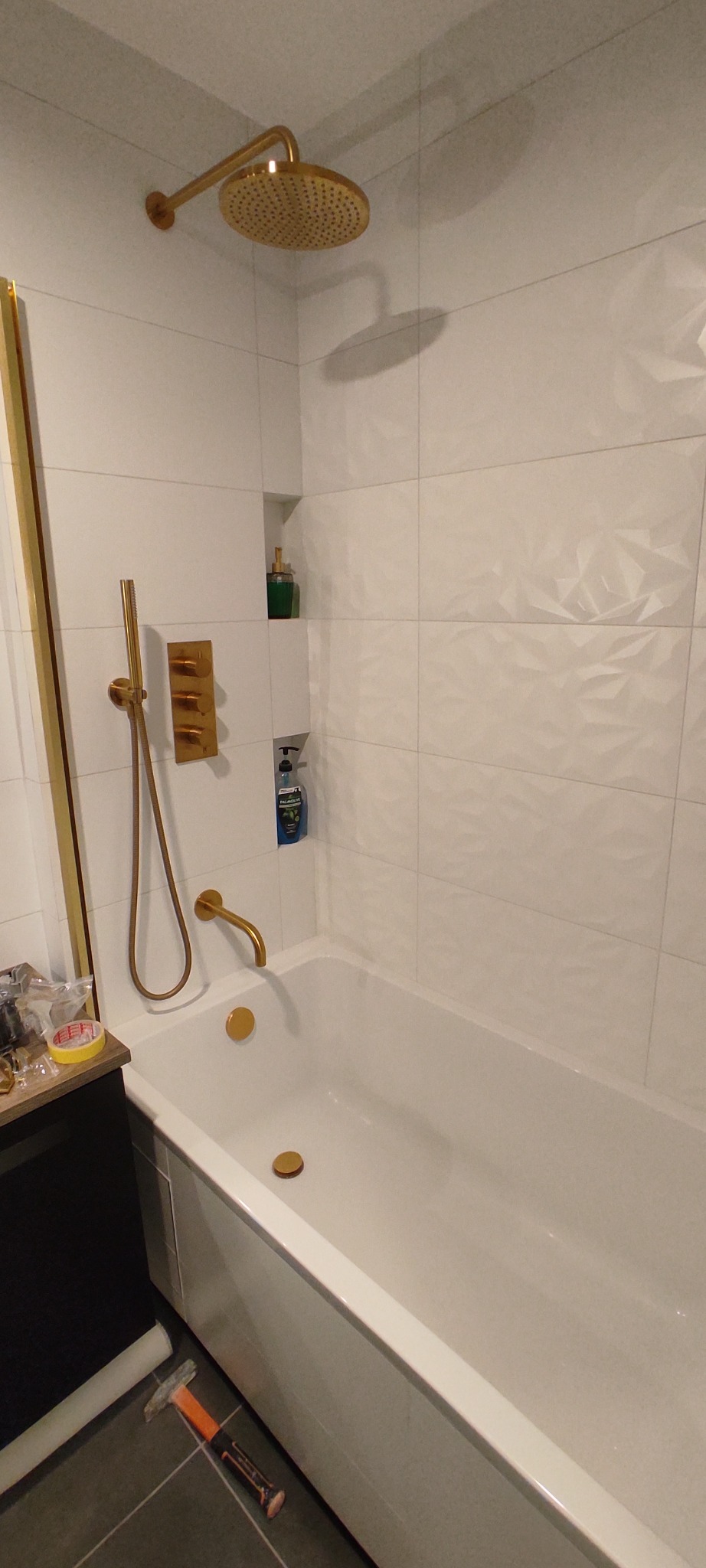 Nowoczesna łazienka z białą wanną, złotą armaturą, deszczownicą i płytkami 3D na ścianie. Na podłodze leży młotek.