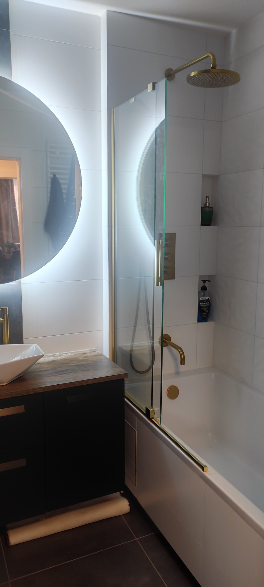 Nowoczesna łazienka z białymi płytkami, okrągłym lustrem z podświetleniem LED, czarną szafką pod umywalką z blatem imitującym drewno i kabiną prysznicową ze złotymi elementami.