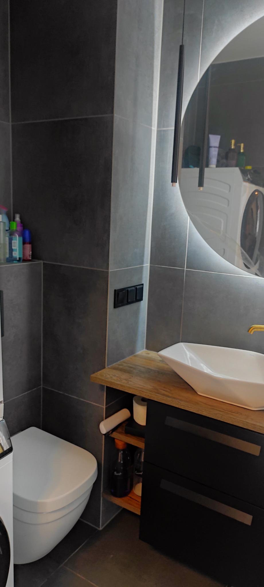 Nowoczesna łazienka z szarymi płytkami, podświetlanym okrągłym lustrem, wiszącą toaletą i umywalką na drewnianym blacie z czarnymi szafkami.