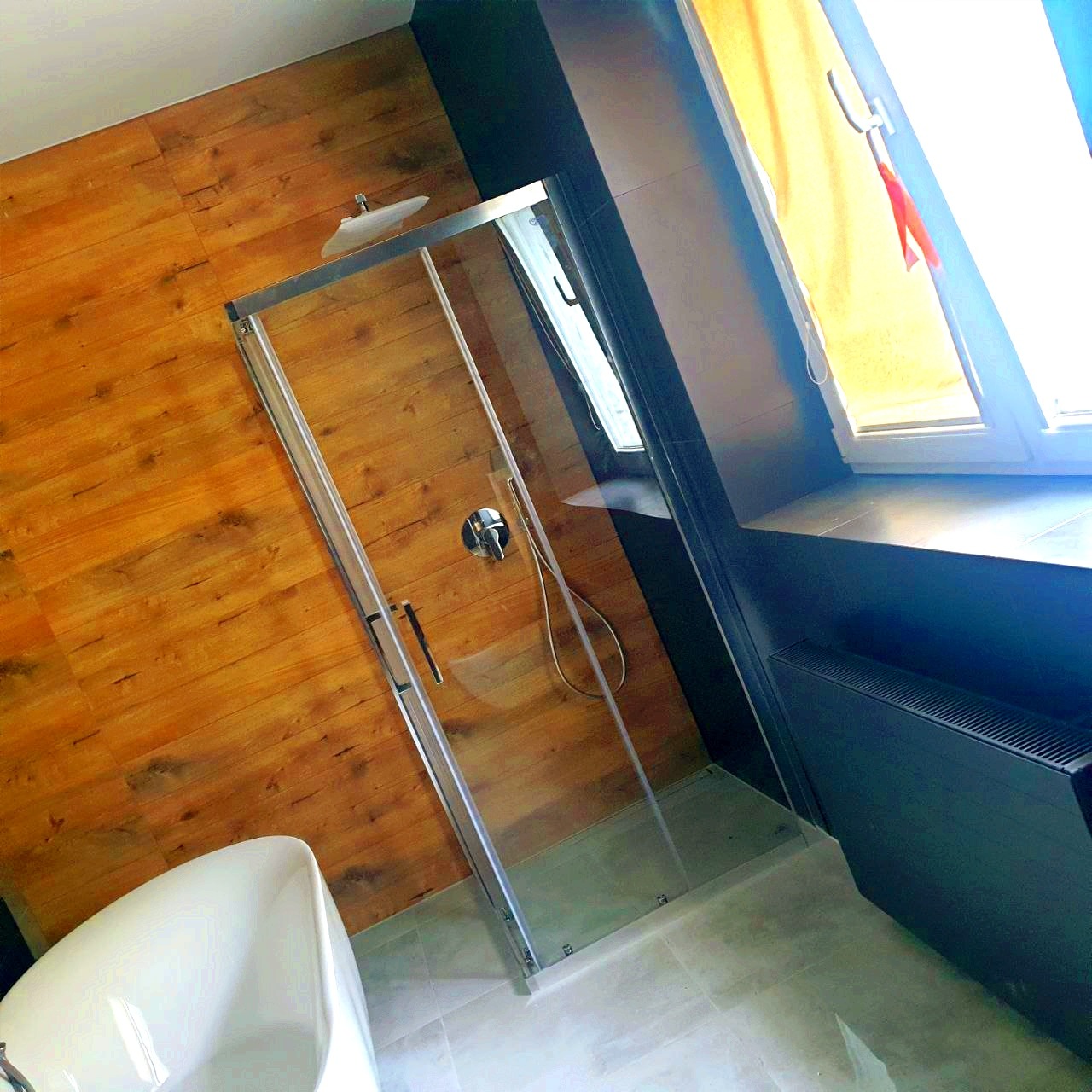 Nowoczesna łazienka z kabiną prysznicową ze szklanymi drzwiami, drewnopodobnymi płytkami na ścianie, białą wanną i ciemną zabudową okna.