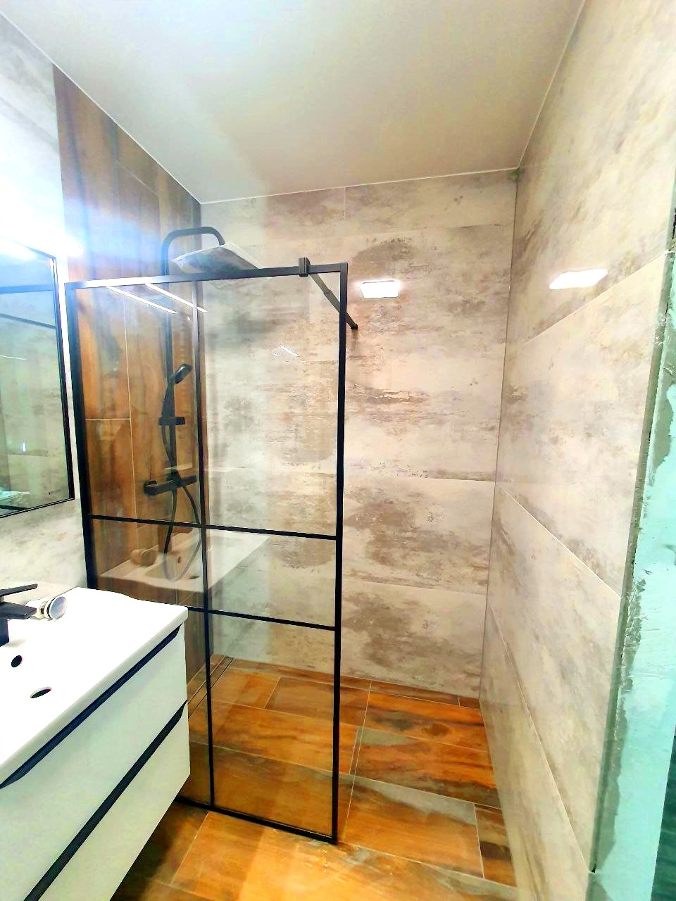 Nowoczesna kabina prysznicowa z czarnymi profilami i szklanymi ściankami w łazience wykończonej płytkami imitującymi drewno, widoczna umywalka z czarną armaturą.