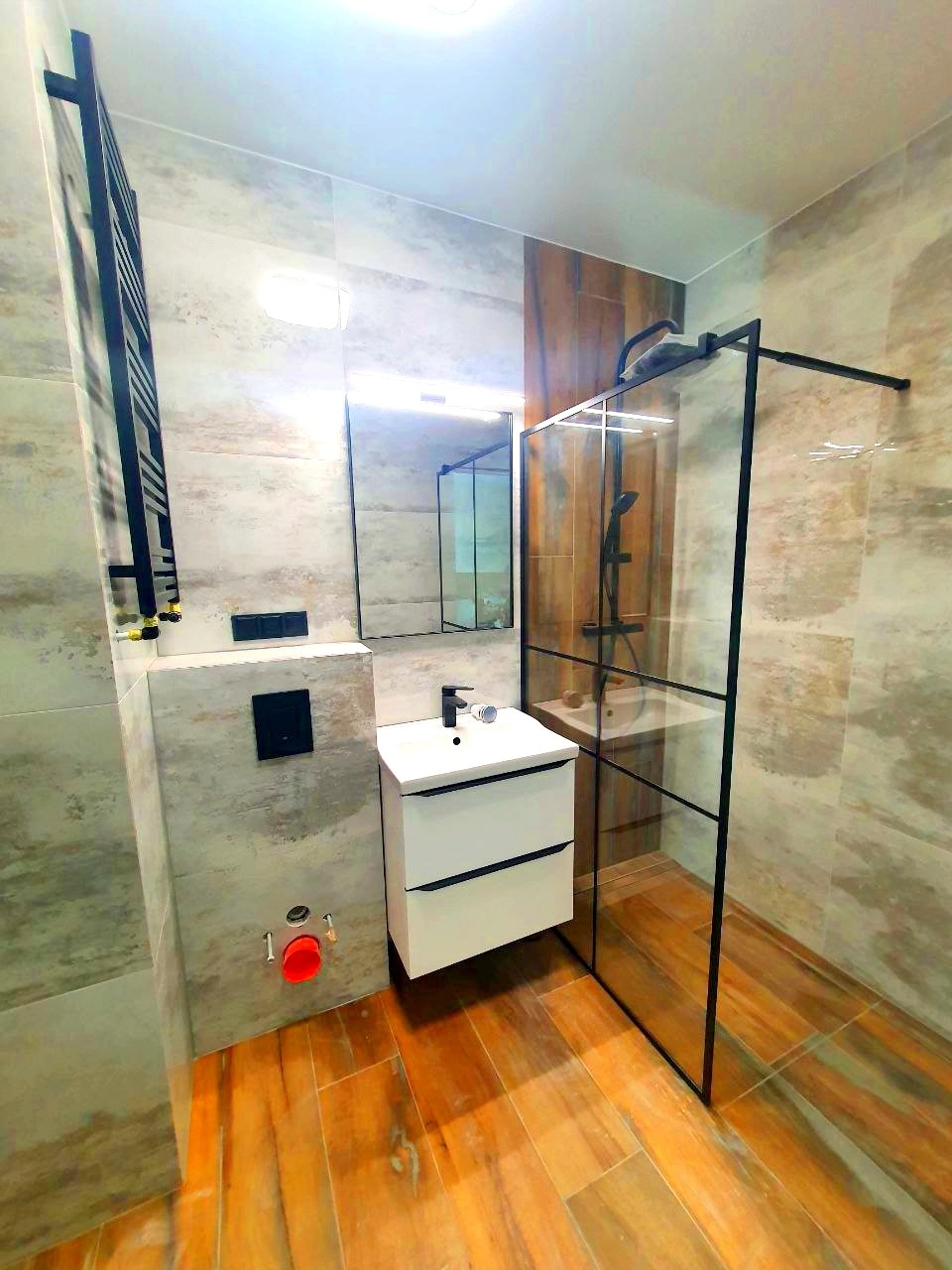 Nowoczesna łazienka z drewnopodobną podłogą i szklaną kabiną prysznicową z czarnymi ramami, biała szafka pod umywalką z czarną armaturą, grzejnik drabinkowy w kolorze czarnym, widoczne przyłącza...