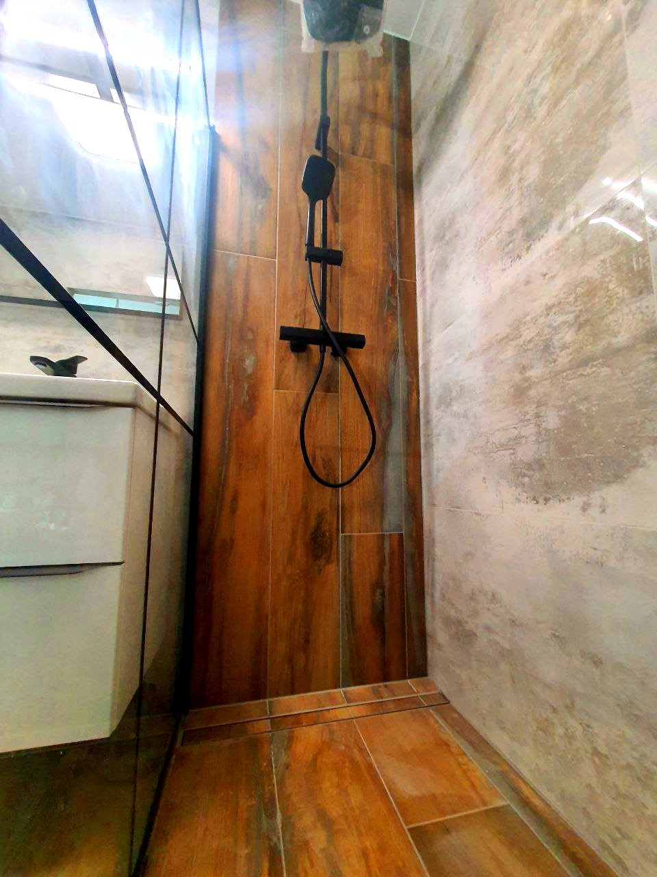 Nowoczesna kabina prysznicowa z drewnopodobnymi płytkami na ścianie i podłodze, czarna armatura, widoczny fragment umywalki i szklana ściana kabiny.