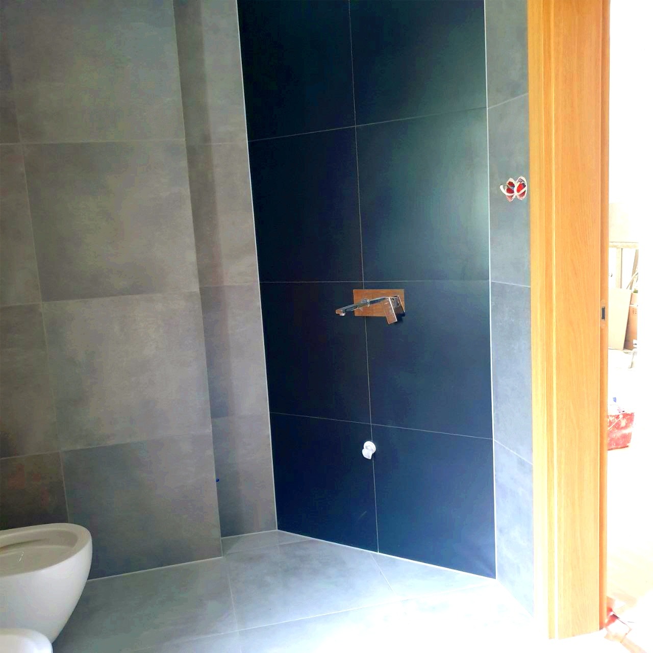 Częściowo wykończona łazienka z szarymi i czarnymi płytkami, widoczna armatura, instalacja elektryczna i fragment toalety, drzwi w ościeżnicy z jasnego drewna.