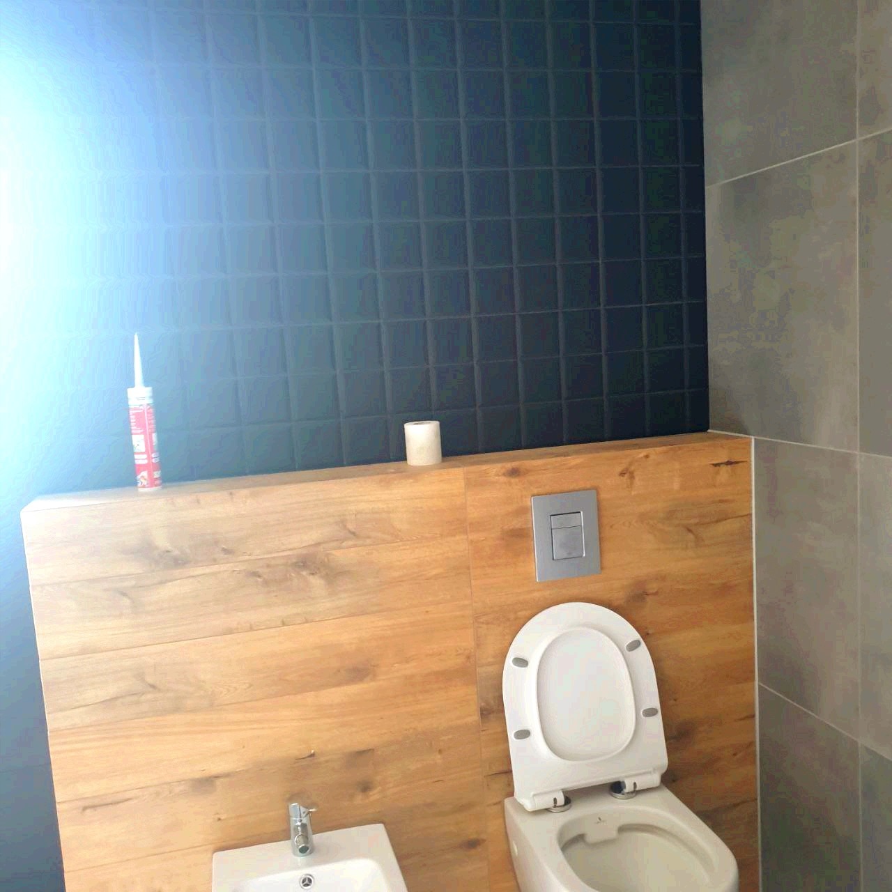Wykończona łazienka z czarnymi płytkami na ścianie, drewnopodobną zabudową, białą toaletą i bidetem; widoczna tuba silikonu i rolka papieru toaletowego.