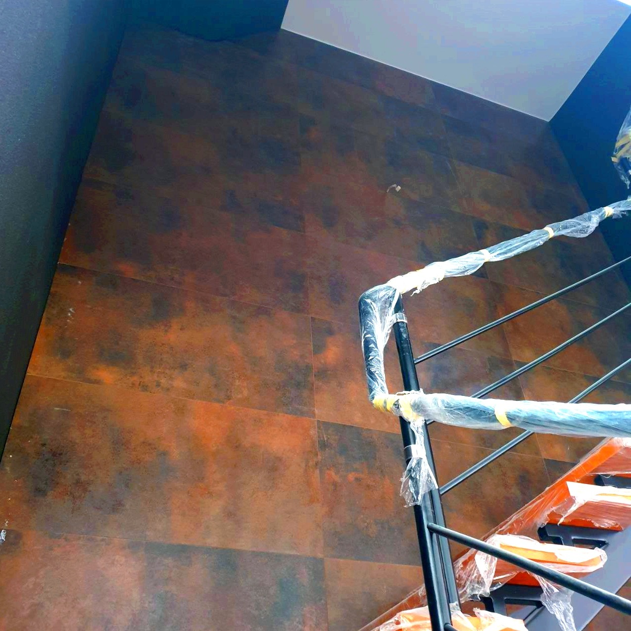 Ściana wyłożona dużymi, brązowo-rdzawymi płytkami ceramicznymi o nieregularnym wzorze, z fragmentem zabezpieczonej folią czarnej metalowej balustrady schodów na pierwszym planie.