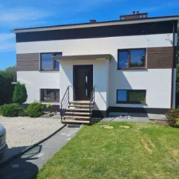 Dwukondygnacyjny dom z płaskim dachem, białymi ścianami i brązowymi akcentami, widok z zewnątrz w słoneczny dzień.