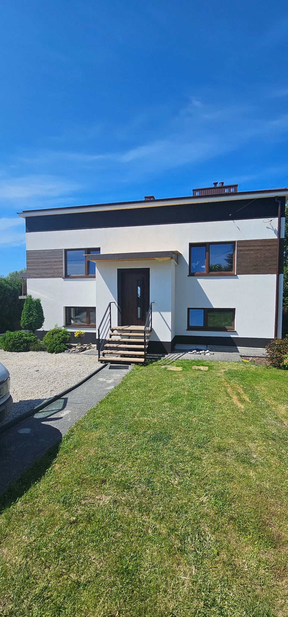 Dwukondygnacyjny dom z płaskim dachem, białymi ścianami i brązowymi akcentami, widok z zewnątrz w słoneczny dzień.