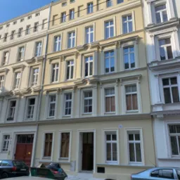 Odnowiona fasada kamienicy w pastelowych kolorach, z nowymi oknami i ozdobnymi elementami architektonicznymi, widok z poziomu ulicy.