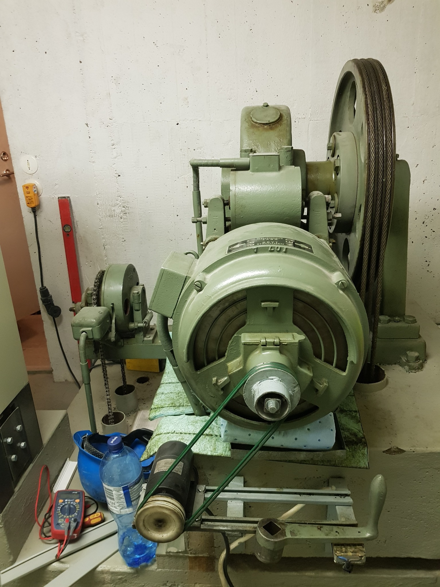 Zielony silnik elektryczny z kołem linowym w maszynowni, obok narzędzia, poziomica i butelka wody.