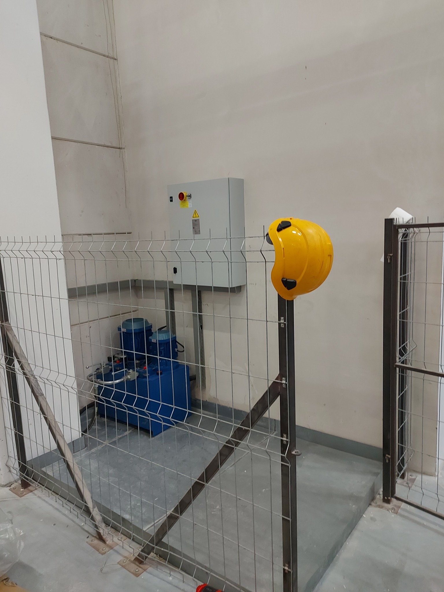 Niebieska hydrauliczna pompa olejowa za metalową siatką, żółty kask na słupku, szara szafka sterownicza z przyciskiem awaryjnym na ścianie w pomieszczeniu technicznym.