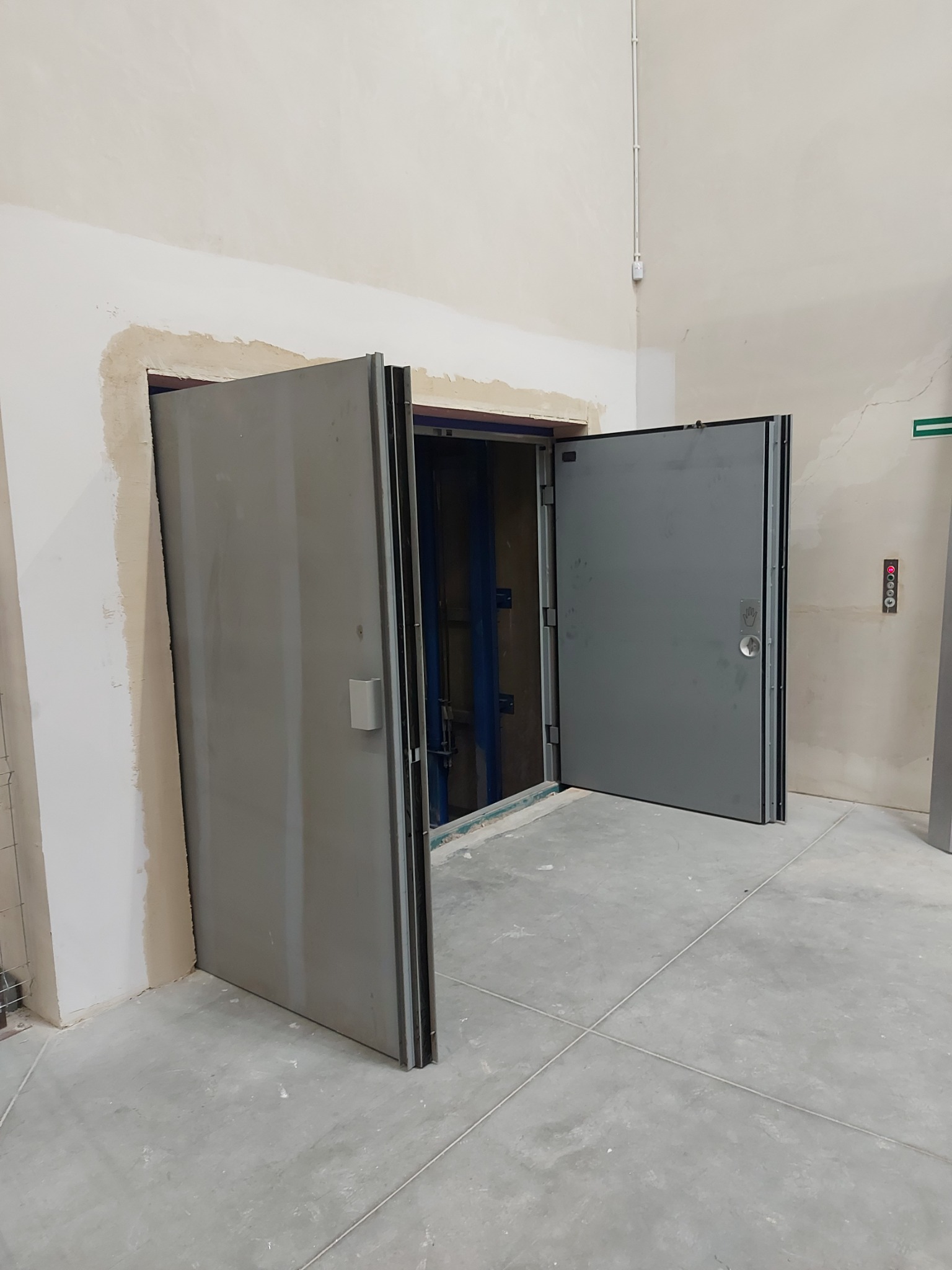 Otwarty, dwuskrzydłowy, szary, metalowy szybowy zamek do windy w stanie surowym, z widoczną konstrukcją szybu, w pomieszczeniu z betonową podłogą i niedokończonymi ścianami.