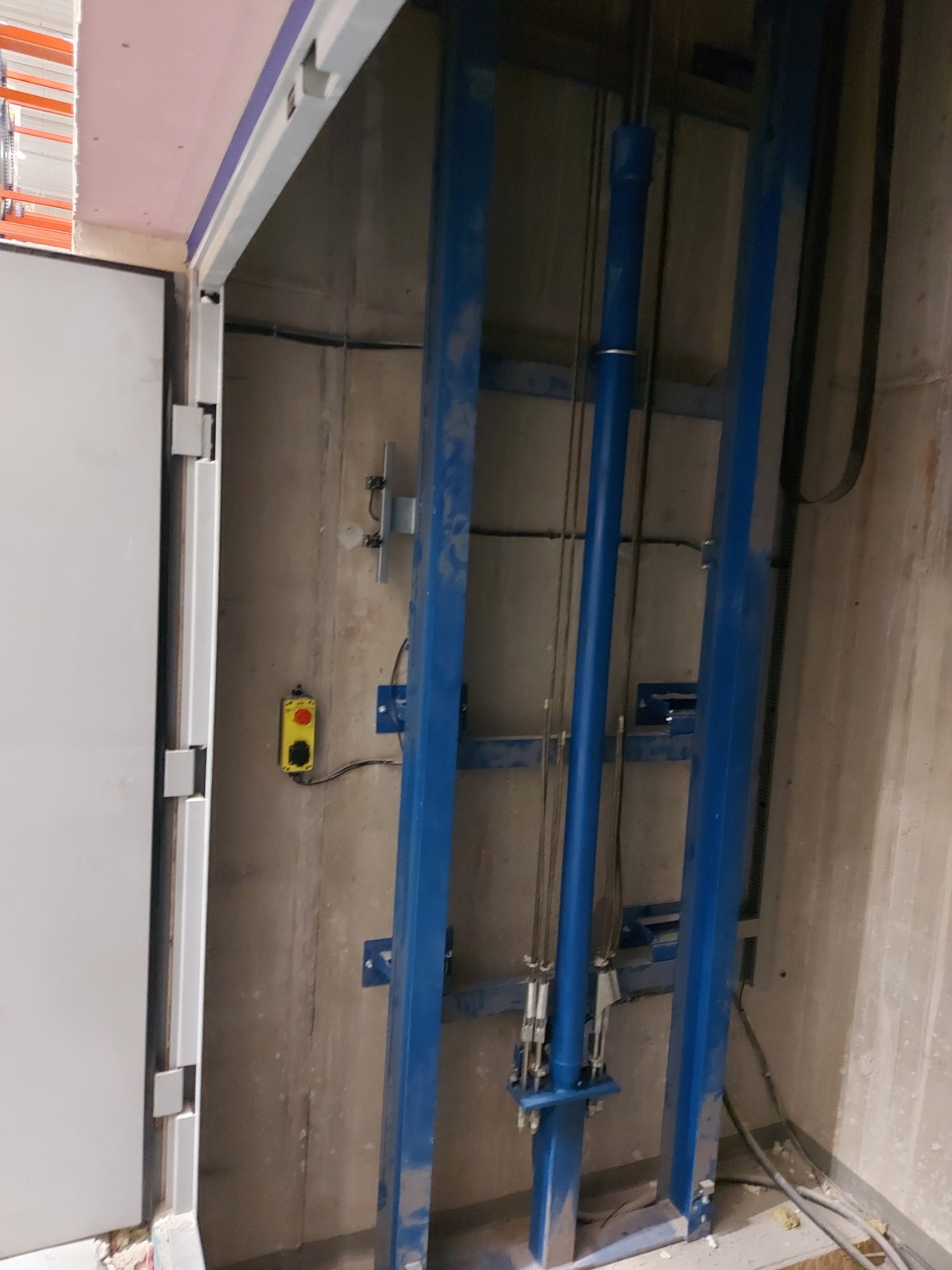 Wnętrze szybu windowego w trakcie montażu, widoczne stalowe elementy konstrukcyjne pomalowane na niebiesko, betonowe ściany, przewody elektryczne i przycisk awaryjny.