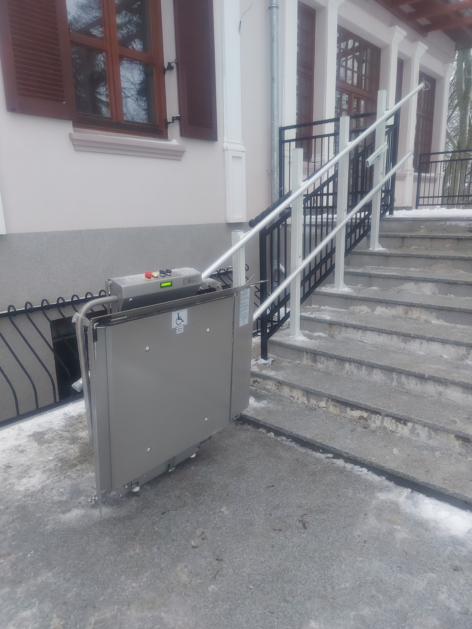 Platforma schodowa dla osób niepełnosprawnych zainstalowana przy wejściu do budynku z widocznymi schodami i barierkami, częściowo pokrytymi śniegiem.