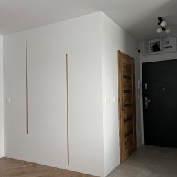 Piotr Karendał - Białe ściany z dekoracyjnymi, pionowymi wstawkami w kolorze mosiądzu, drewniana wewnętrzna ościeżnica z czarnymi akcentami, czarne drzwi wejściowe, parkiet w jodełkę i szara podłoga w przedpokoju.