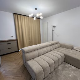 Piotr Karendał - Beżowy, modułowy narożnik z tkaniny o prążkowanej strukturze, stojący na szarym dywanie ze wzorem, w tle komoda, zasłony i lampa sufitowa w nowoczesnym salonie z parkietem w jodełkę.