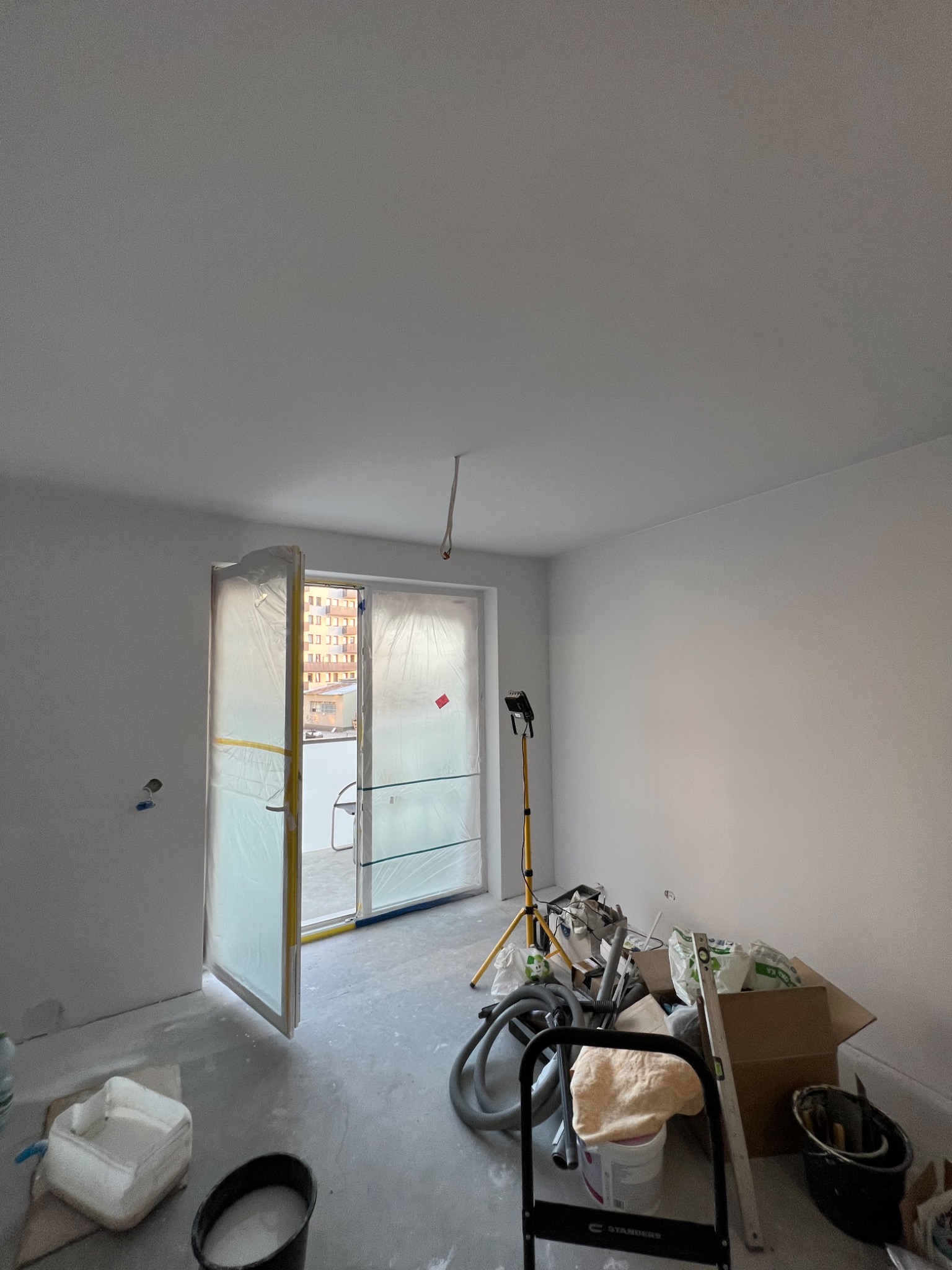 Pokój w trakcie malowania na biało, okno zabezpieczone folią malarską, widoczne narzędzia malarskie i oświetlenie budowlane.