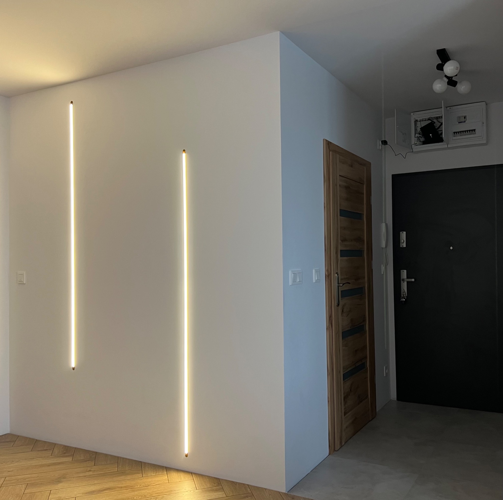Nowoczesne oświetlenie ścienne w przedpokoju: dwie pionowe lampy LED na białej ścianie, parkiet ułożony w jodełkę na podłodze, widoczna drewniana ościeżnica drzwi wewnętrznych i czarne drzwi...