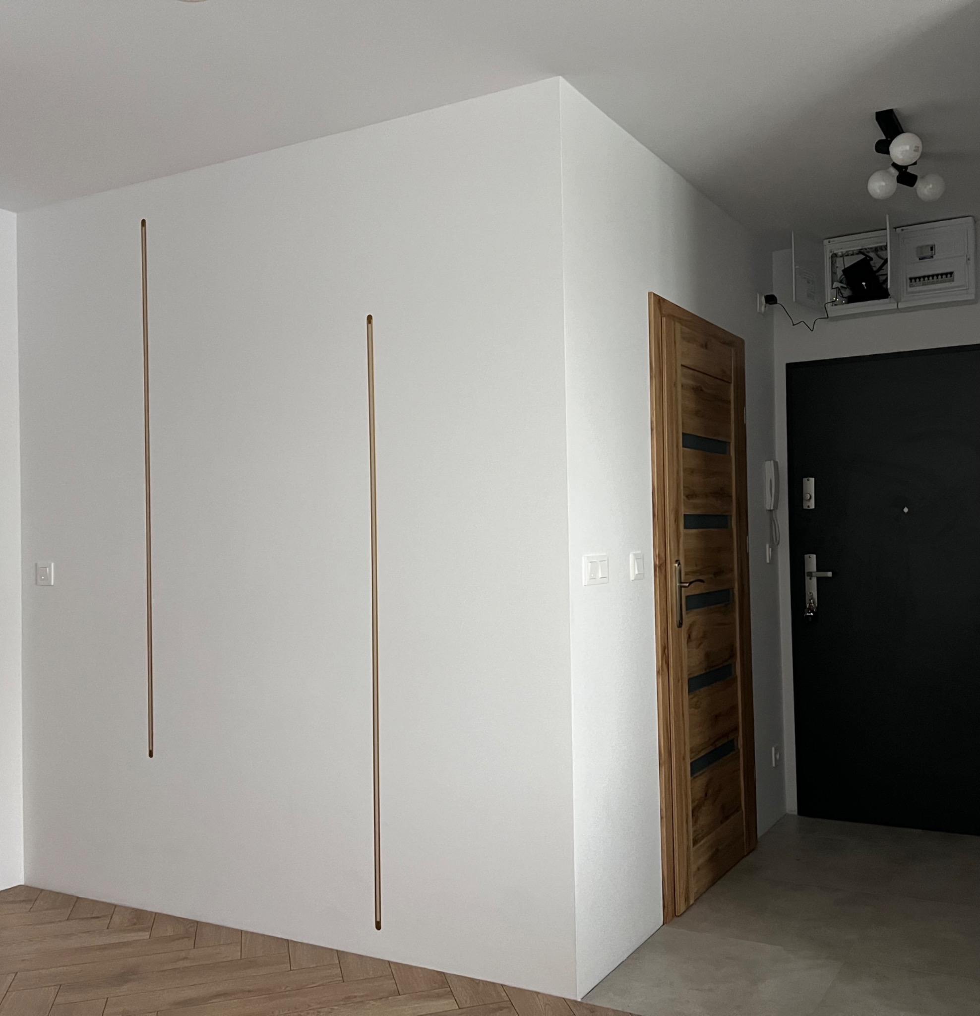 Białe ściany z dekoracyjnymi, pionowymi wstawkami w kolorze mosiądzu, drewniana wewnętrzna ościeżnica z czarnymi akcentami, czarne drzwi wejściowe, parkiet w jodełkę i szara podłoga w przedpokoju.