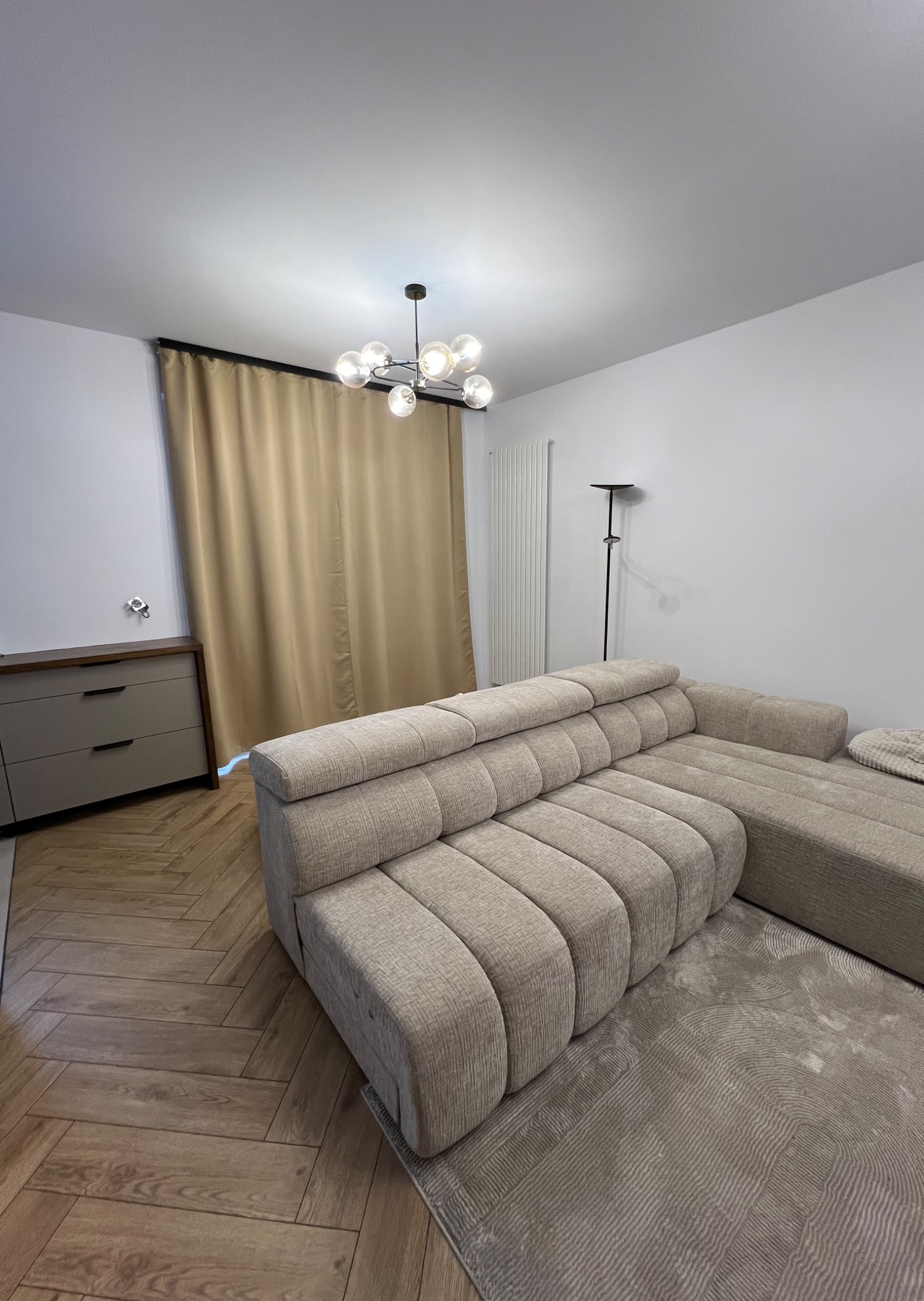 Beżowy, modułowy narożnik z tkaniny o prążkowanej strukturze, stojący na szarym dywanie ze wzorem, w tle komoda, zasłony i lampa sufitowa w nowoczesnym salonie z parkietem w jodełkę.