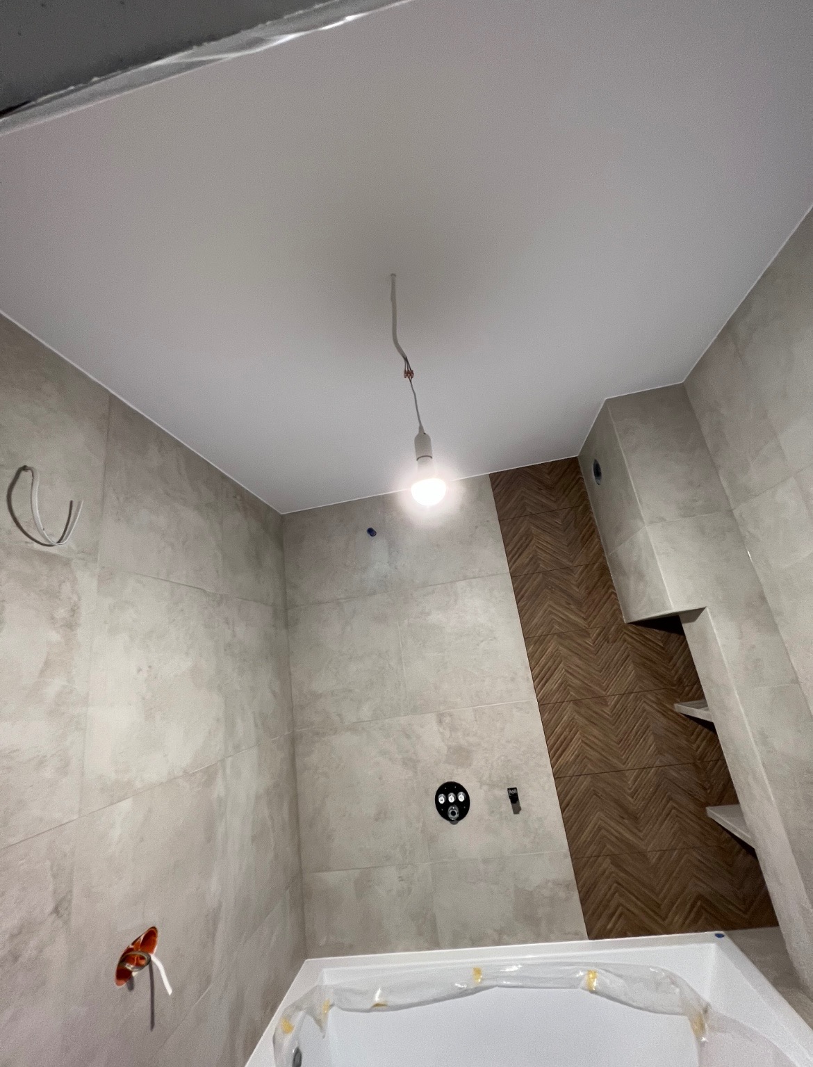 Łazienka w trakcie wykańczania: jasne płytki na ścianach, biały sufit napinany z wiszącą żarówką, dekoracyjna wstawka z drewnopodobnych płytek ułożonych w jodełkę, widoczna część wanny...