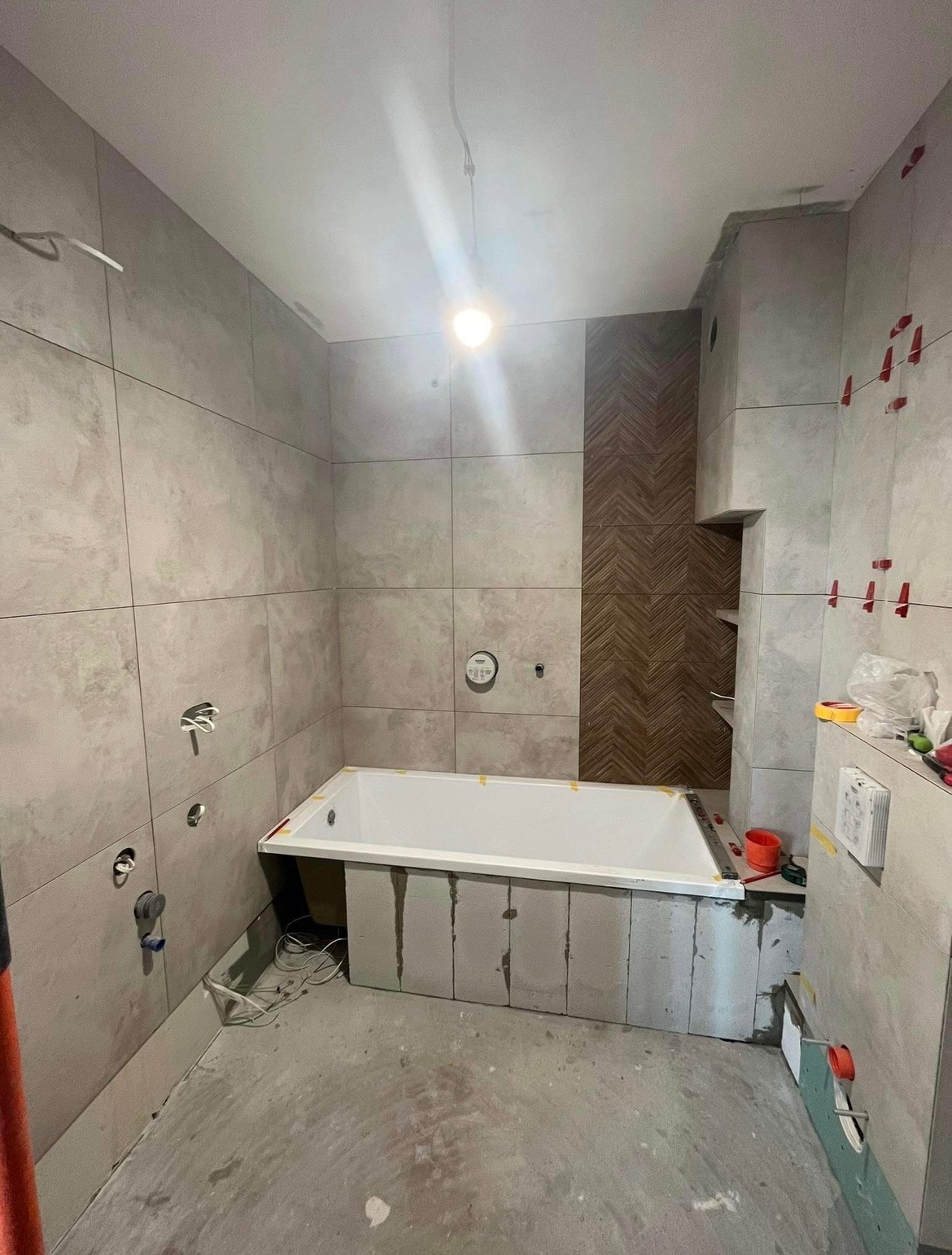 Łazienka w trakcie prac wykończeniowych: biała wanna obudowana bloczkami, szare płytki na ścianach z czerwonymi krzyżykami dystansowymi, dekoracyjny panel z drewnopodobnych płytek ułożonych...