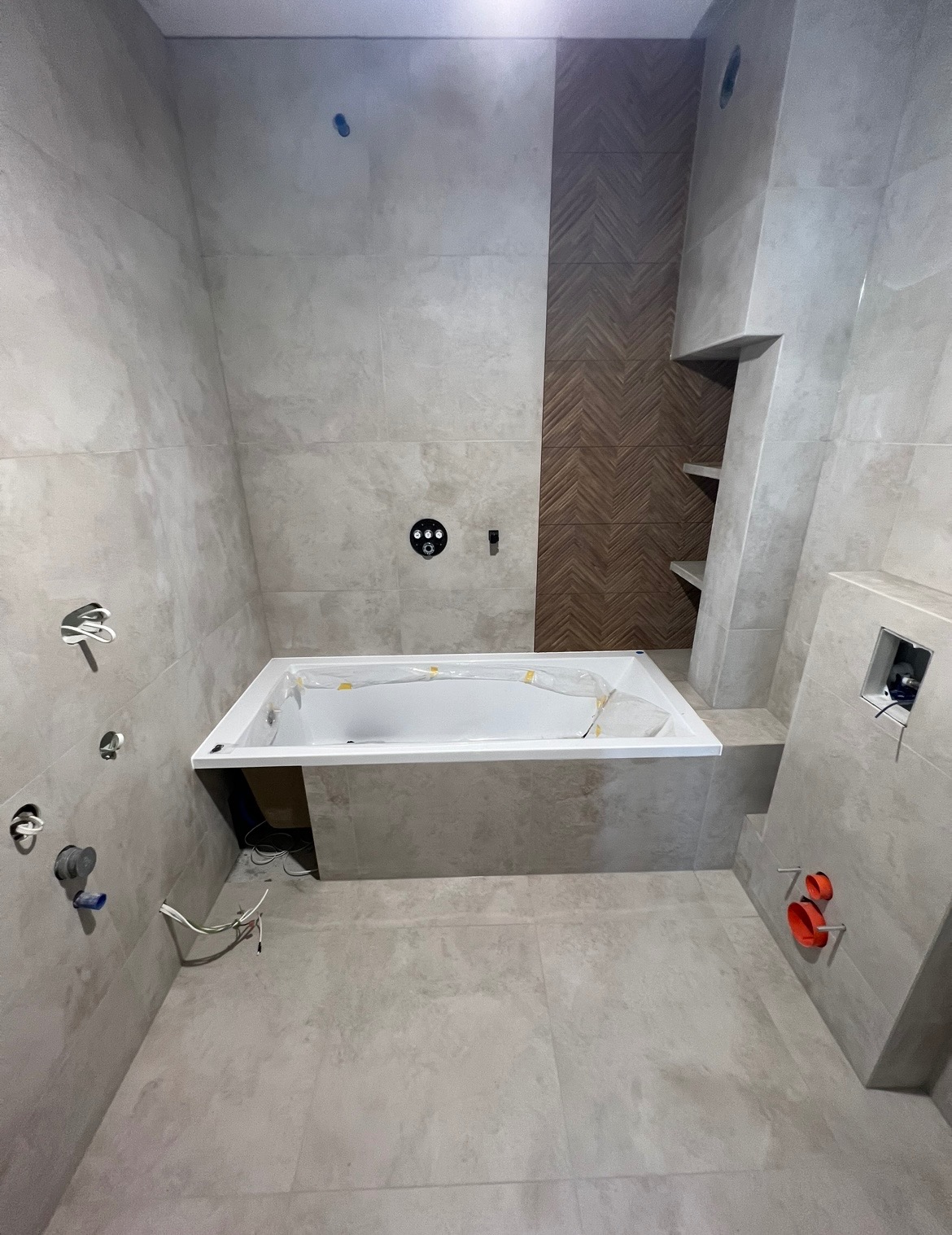 Nowoczesna łazienka w trakcie remontu: biała wanna zabezpieczona folią, szare płytki imitujące kamień, drewnopodobna mozaika w pionowym pasie, widoczne instalacje wodne i elektryczne przed montażem...