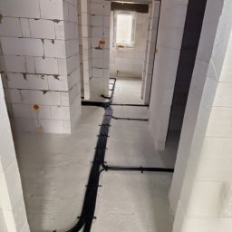 Instal-Lux Instalacje elektryczne Paweł Mizerski - Wnętrze budynku w trakcie budowy z widocznymi kablami elektrycznymi prowadzonymi po podłodze i ścianach, przymocowanymi uchwytami, ściany z białych bloczków, surowy stan wykończenia.