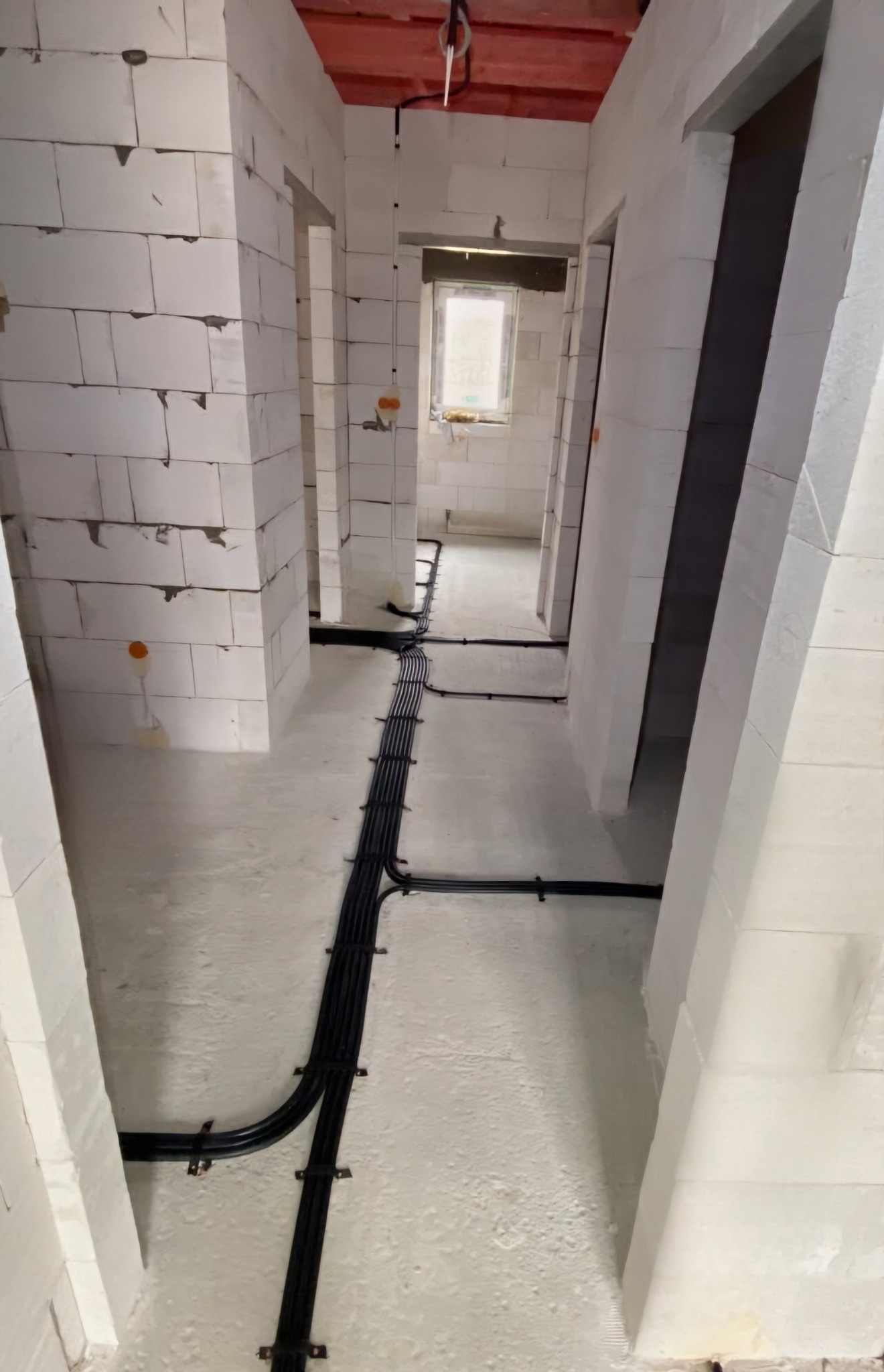 Wnętrze budynku w trakcie budowy z widocznymi kablami elektrycznymi prowadzonymi po podłodze i ścianach, przymocowanymi uchwytami, ściany z białych bloczków, surowy stan wykończenia.