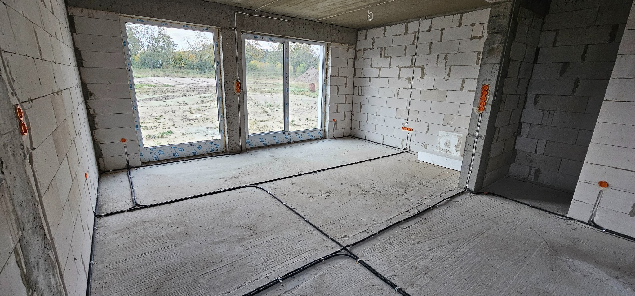 Surowy stan wnętrza z widocznymi przewodami elektrycznymi ułożonymi na posadzce, pomarańczowe puszki elektryczne w ścianach z betonu komórkowego, dwa okna z widokiem na zewnątrz.