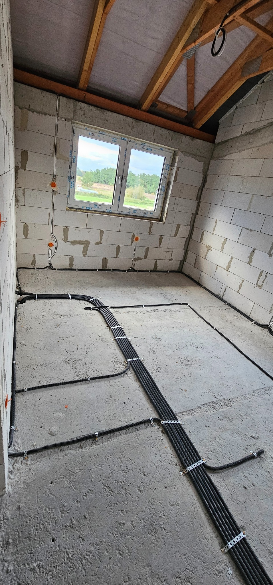 Surowy stan poddasza z widoczną instalacją elektryczną w peszlach, przymocowaną do betonowej posadzki i ścian z bloków, okablowanie prowadzone pod oknem z widokiem na zieleń.