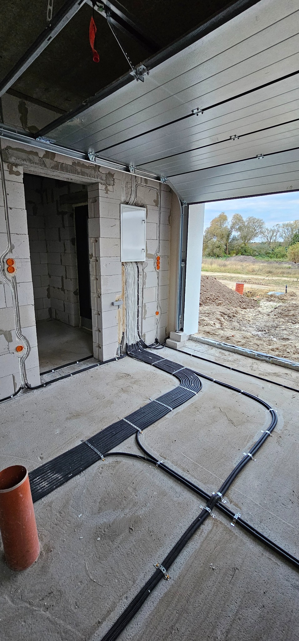 Wnętrze garażu w trakcie budowy z widocznymi przewodami elektrycznymi ułożonymi w korytach na podłodze i wyprowadzonymi do skrzynki na ścianie z pustaków. Widoczne pomarańczowe puszki pod gniazdka.