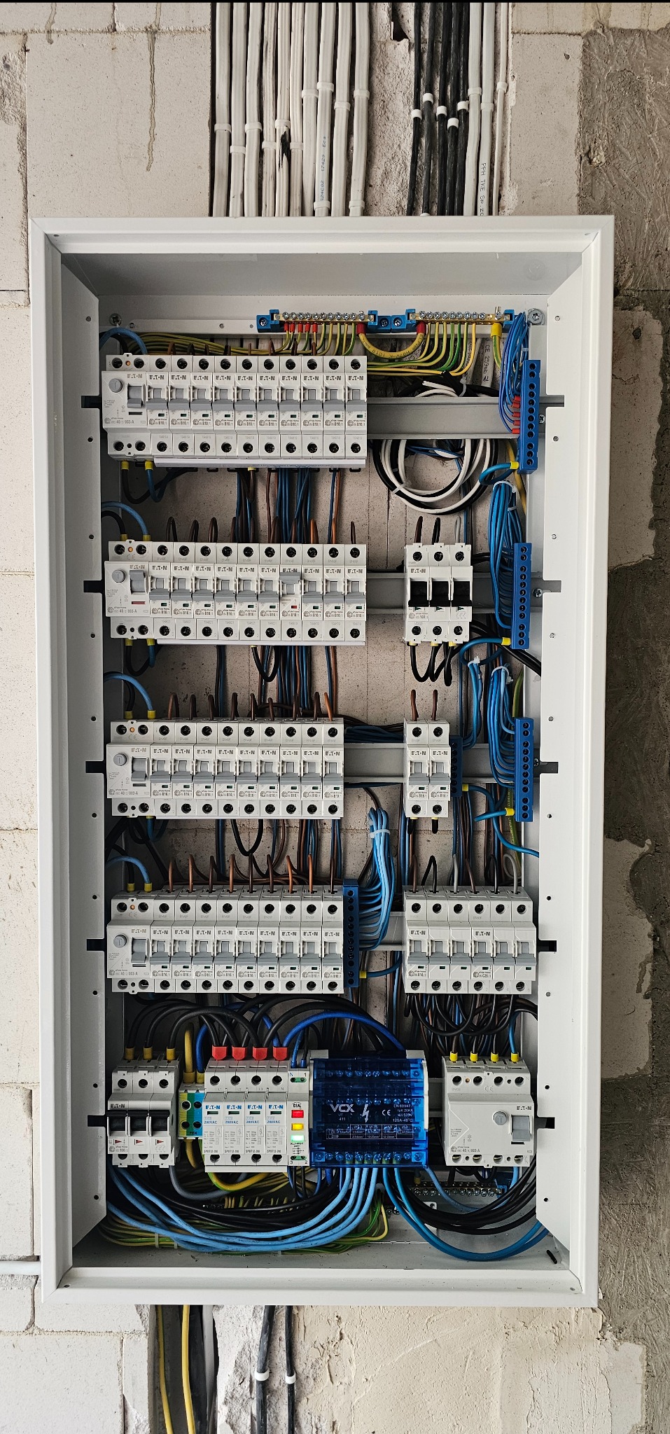 Rozdzielnica elektryczna z zabezpieczeniami nadprądowymi i przeciwprzepięciowymi, z widocznymi przewodami w kolorach żółtym, niebieskim i brązowym, zamontowana na surowej ścianie z betonu komórkowego.