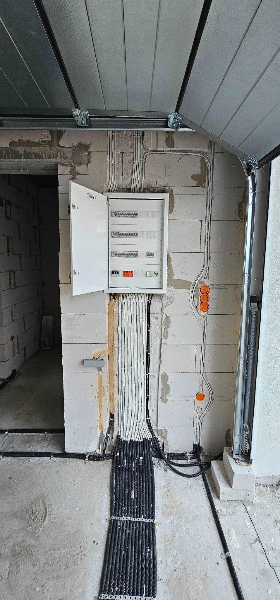 Otwarta skrzynka z bezpiecznikami zamontowana na ścianie z betonu komórkowego. Widoczne liczne przewody elektryczne prowadzone w peszlach, zarówno w ścianie, jak i na jej powierzchni. Montaż...