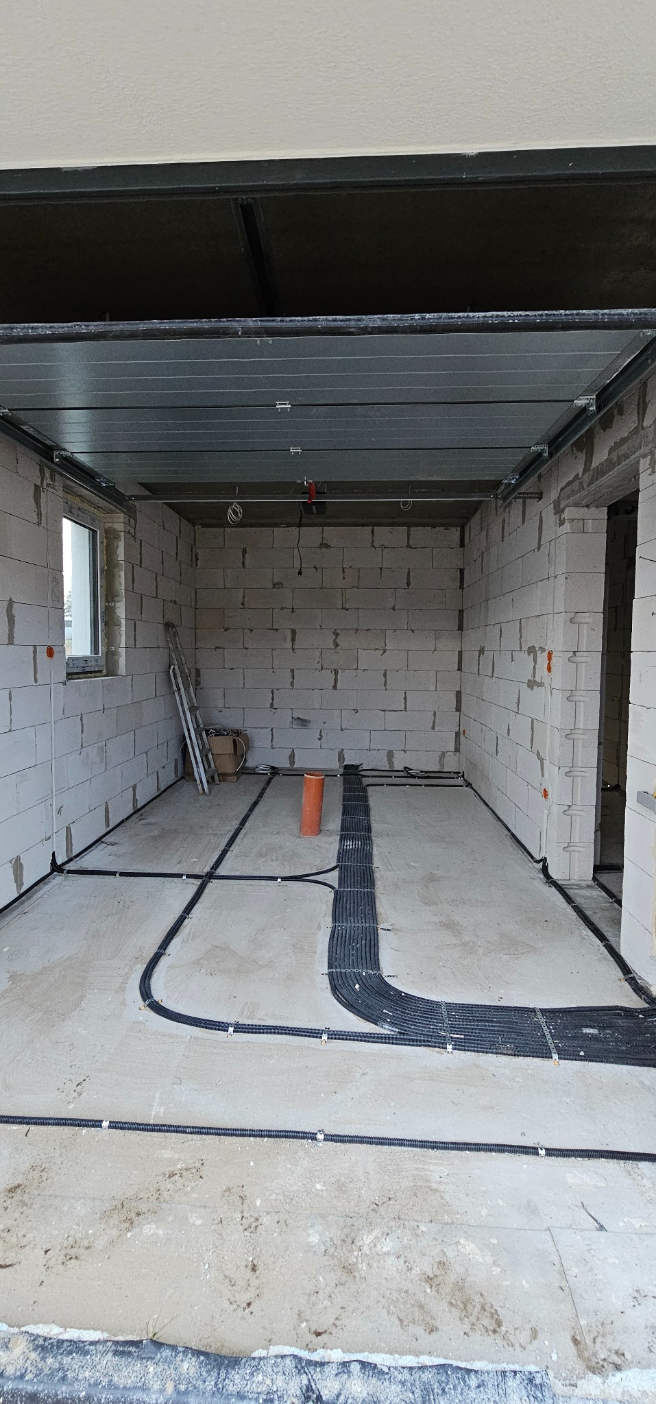Wnętrze surowego garażu z widoczną instalacją elektryczną w peszlach ułożoną na posadzce, drabina oparta o ścianę z bloczków.