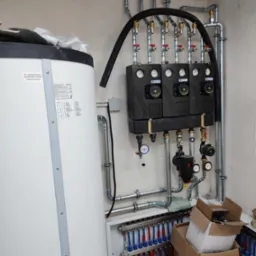 Biały zasobnik ciepłej wody użytkowej, rozdzielacz ogrzewania podłogowego z niebiesko-czerwonymi pętlami oraz zestaw pompowo-mieszający z trzema pompami obiegowymi i manometrami, instalacja...