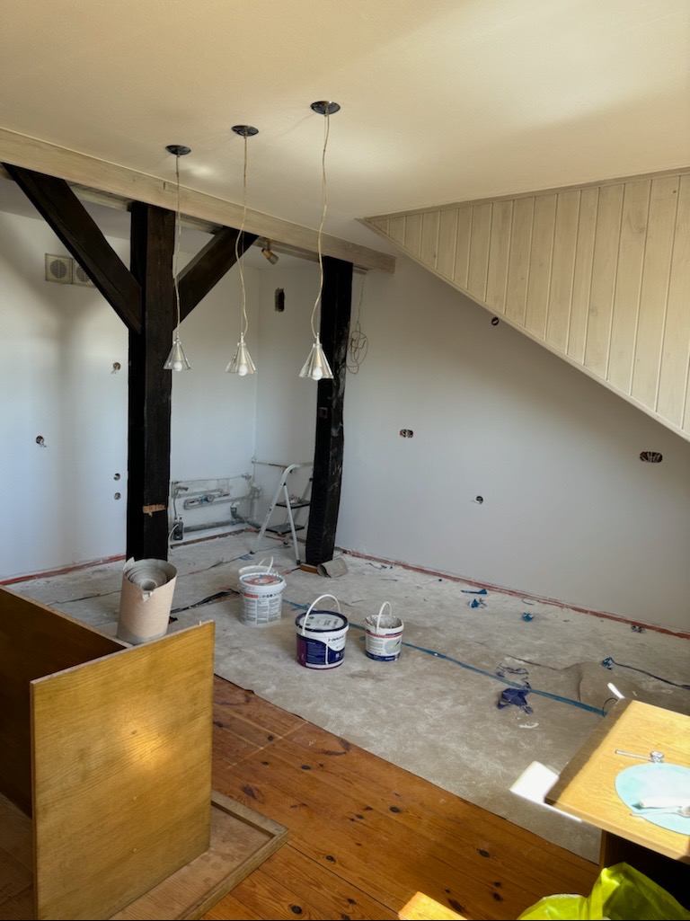 Poddasze w trakcie remontu, ściany świeżo pomalowane na biało, widoczne drewniane elementy konstrukcyjne, trzy wiszące lampy, podłoga zabezpieczona folią malarską, kilka wiader z farbą i drabina.