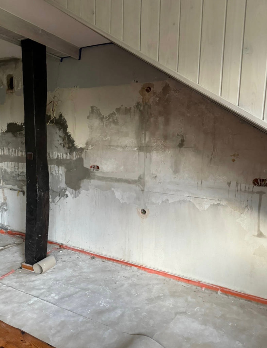 Ściana w trakcie przygotowania do prac wykończeniowych, widoczne ślady po instalacjach, zabezpieczona podłoga, drewniany słup i fragment boazerii na poddaszu.