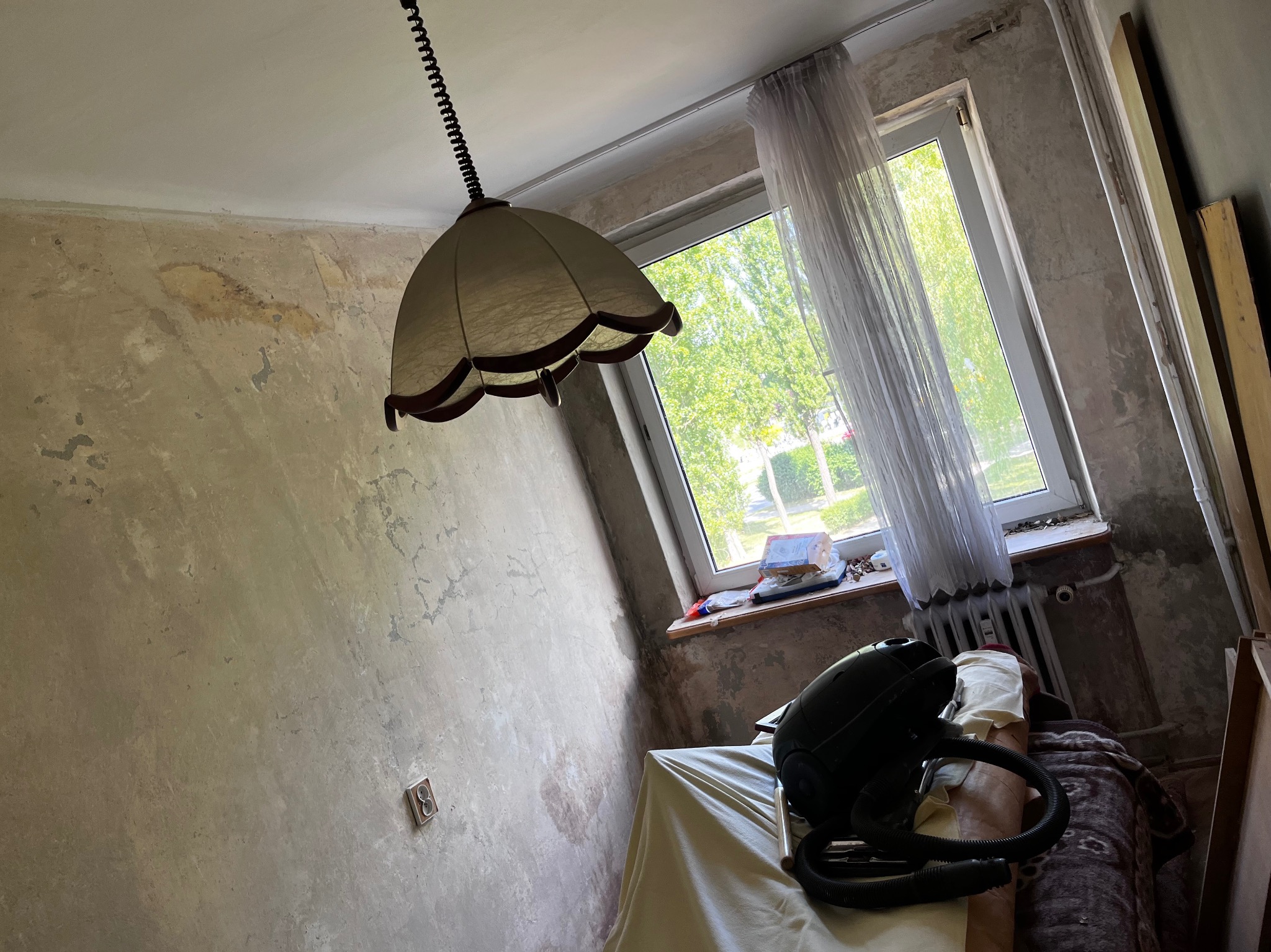 Pokój w trakcie remontu: ściany z widocznymi śladami starej farby, wisząca lampa retro i odkurzacz stojący na przykrytym meblu, okno z firanką w tle.