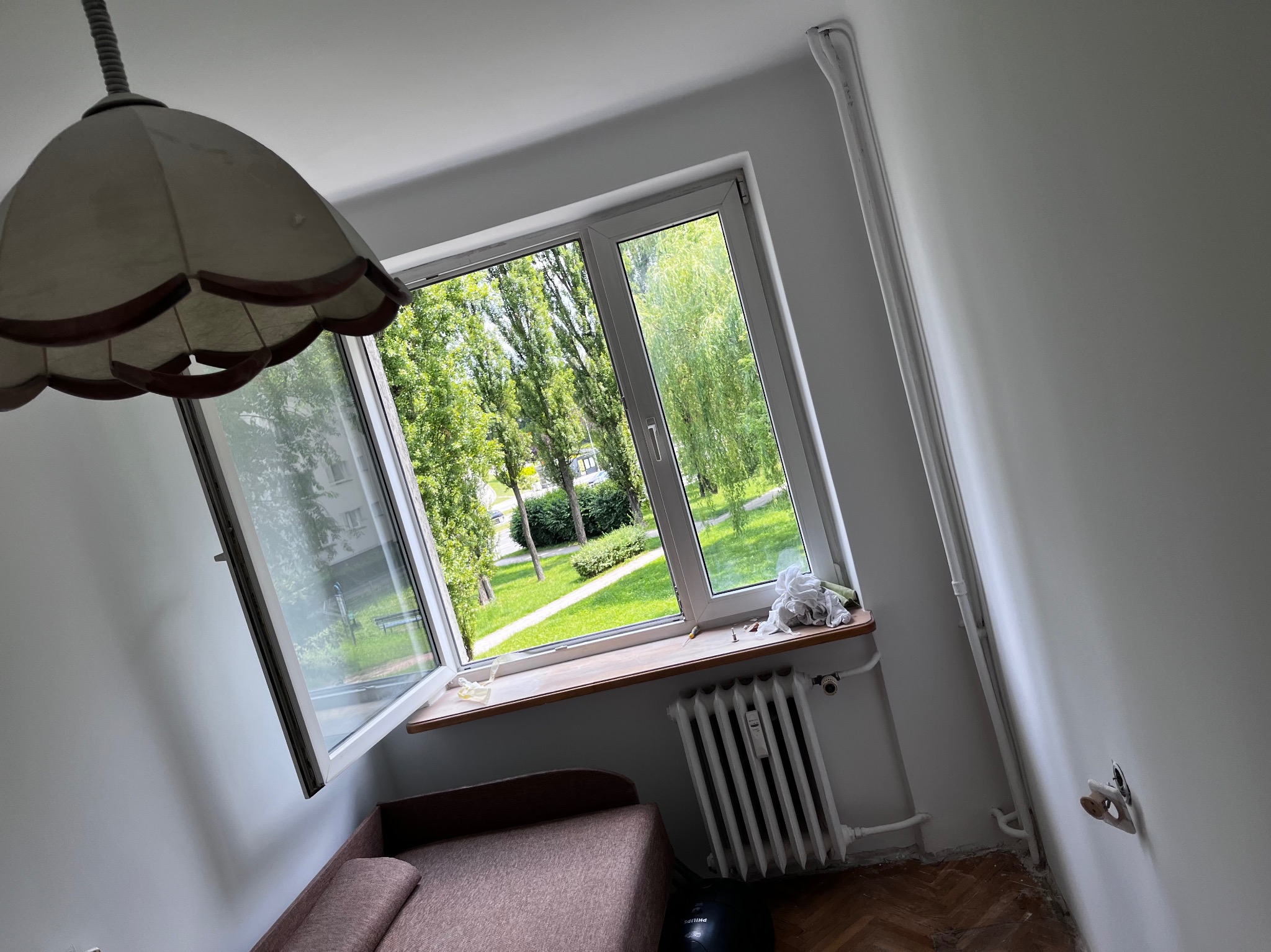 Wnętrze pokoju po odświeżeniu, z otwartym oknem wychodzącym na zieleń; widoczny odnowiony grzejnik oraz świeżo pomalowane ściany w jasnym odcieniu.