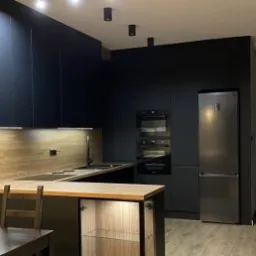 Nowoczesna kuchnia z czarnymi matowymi szafkami, drewnianym blatem i oświetleniem punktowym, widok na stół z krzesłami i fragment sąsiedniego pomieszczenia.