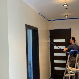 BajaHaus - Mężczyzna maluje ścianę w korytarzu wałkiem na teleskopowym kiju, stojąc na drabinie; widoczne taśmy malarskie zabezpieczające sufit i futryny, surowe lampy sufitowe bez kloszy.