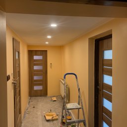 BajaHaus - Korytarz w trakcie odświeżania, ściany pomalowane na ciepły odcień brzoskwini, widoczna drabina malarska z akcesoriami, zabezpieczona podłoga folią malarską, trzy drewniane drzwi z przeszkleniami.