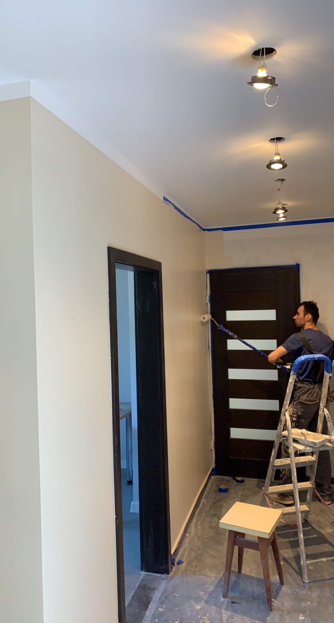 Mężczyzna maluje ścianę w korytarzu wałkiem na teleskopowym kiju, stojąc na drabinie; widoczne taśmy malarskie zabezpieczające sufit i futryny, surowe lampy sufitowe bez kloszy.