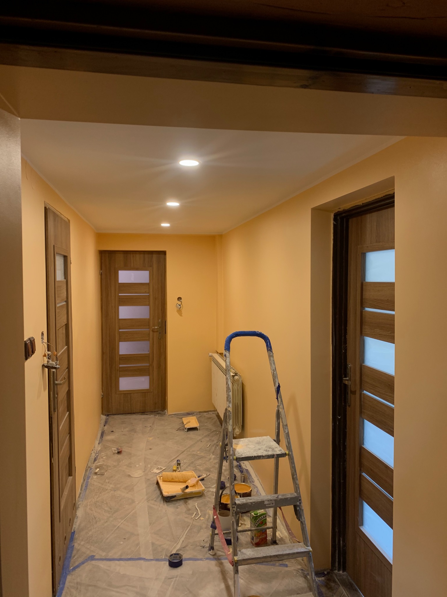 Korytarz w trakcie odświeżania, ściany pomalowane na ciepły odcień brzoskwini, widoczna drabina malarska z akcesoriami, zabezpieczona podłoga folią malarską, trzy drewniane drzwi z przeszkleniami.
