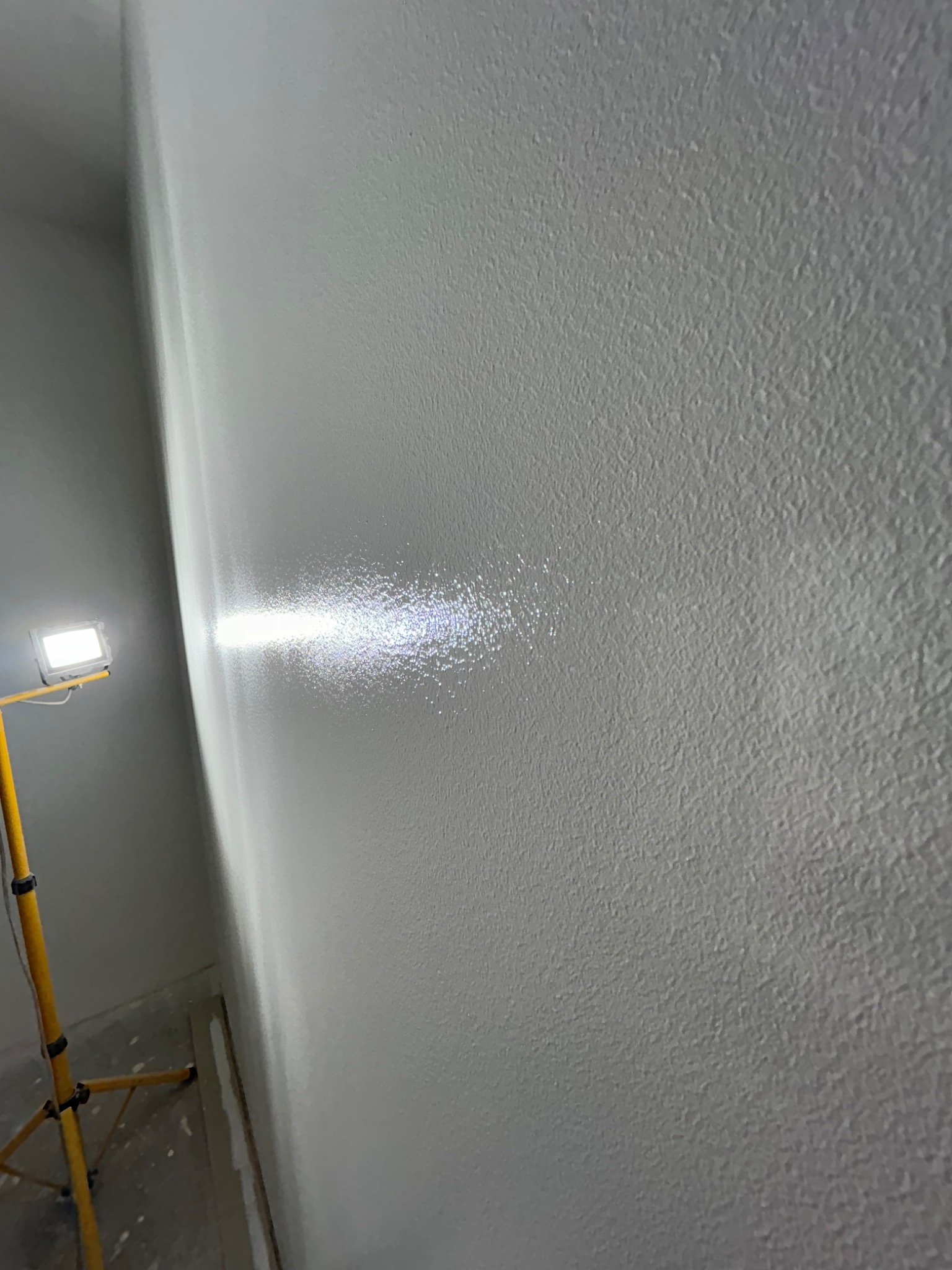 Świeżo pomalowana, jasnoszara ściana o wyraźnej fakturze oświetlona lampą roboczą, widoczny fragment podłogi w trakcie prac.
